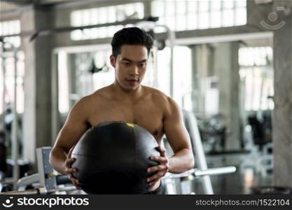 Athletic Asian muscle man with nake torso workout on abdomen ball at fitness gym. Man exercise by swinging hard medicine ball in sport club. Bodybuilding and healthy lifestyle concept.