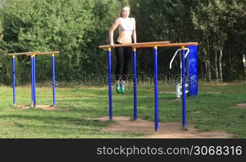 Athletic girl exercising on parallel bars on sports ground on bright summer day.