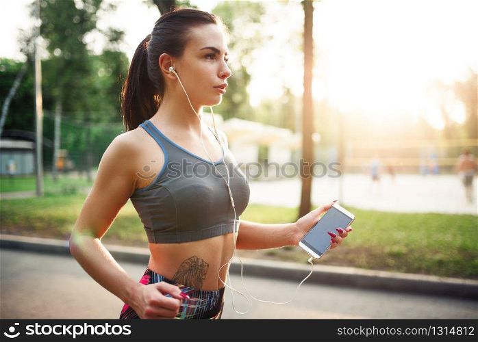 Athletic girl jogging with headphones on walkway in summer park. Woman on outdoors morning workout. Athletic girl jogging with headphones in park