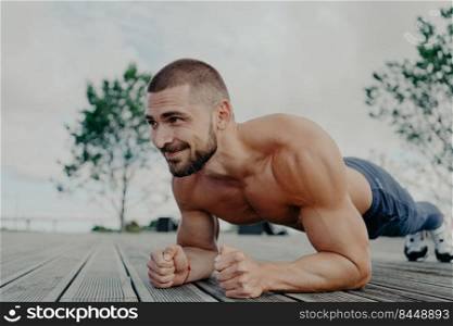 Athletic guy stands in plank pose to keep fit, leads healthy lifestyle and poses outdoor. Young bearded man in good physical shape does abdominal excersice, has muscular torso. Bodybuilding concept