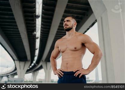 Athletic male fitness model keeps hands on waist poses with muscular perfect naked body, looks seriously into distance, does sport exercises outdoor, focused somewhere, goes in for sport regularly