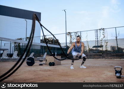 Athletic man doing exercise with ropes, street workout, crossfit. Fitness training on sports ground outdoor, male person pumps muscles, active urban lifestyle. Athletic man with ropes, street workout, crossfit