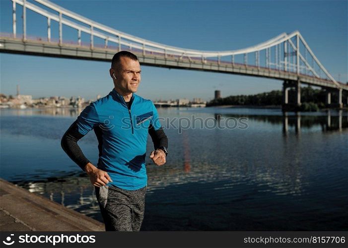 Athletic man doing morning training workout and running on river bank. City bridge on background. Athletic man running on river bank over city bridge on background