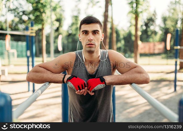 Athletic man in headphones doing exercise on parallel bars, front view, outdoor fitness workout. Strong sportsman on sport training in park. Man doing exercise on parallel bars, front view