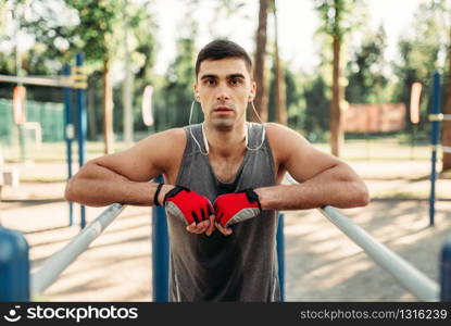 Athletic man in headphones doing exercise on parallel bars, front view, outdoor fitness workout. Strong sportsman on sport training in park. Man doing exercise on parallel bars, front view