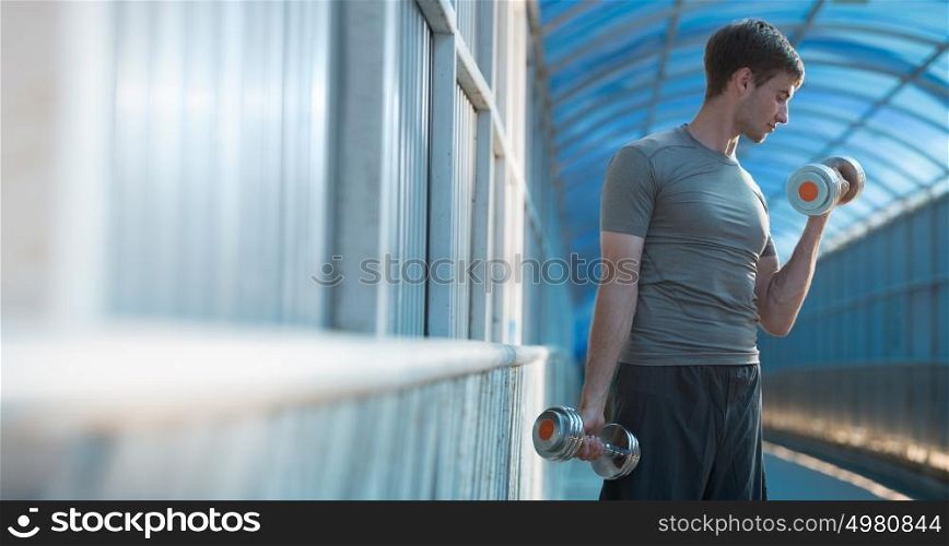 Athletic man pumping up muscles with dumbbells outdoors in city