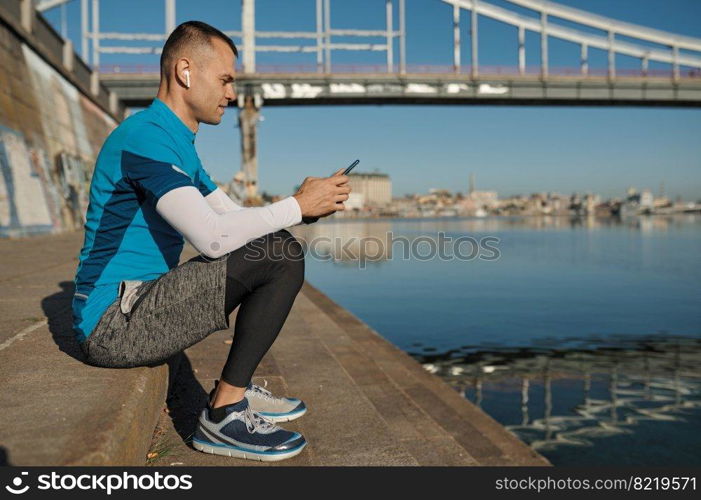 Athletic man rest with smartphone after running functional workout outdoors. Sportsman listening to music, checking health indicator after jogging on city river bank. Athletic man rest after running functional workout outdoors