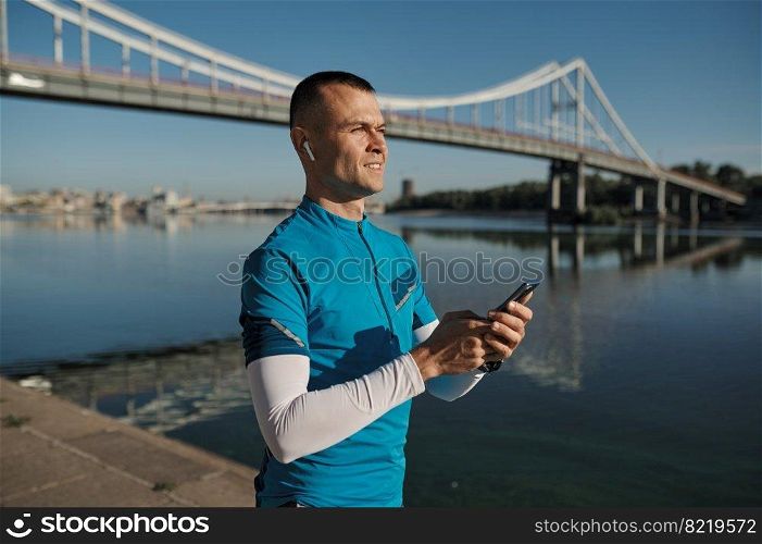 Athletic man rest with smartphone after running functional workout outdoors. Sportsman listening to music, checking health indicator after jogging on city river bank. Athletic man rest after running functional workout outdoors