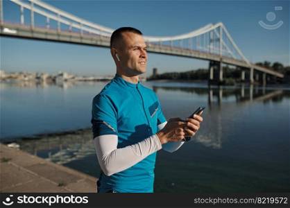 Athletic man rest with smartphone after running functional workout outdoors. Sportsman listening to music, checking health indicator after jogging on city river bank. Athletic man rest after running functional workout outdoors