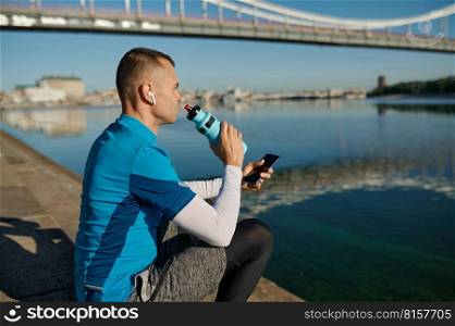 Athletic man rest with smartphone and bottle of water after running functional workout outdoors. Sportsman listening to music, checking health indicator after jogging on city river bank. Athletic man rest after running functional workout outdoors