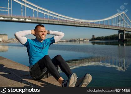 Athletic man runner building abs muscles lying on ground. Sportsman warming up outdoors in summer day. Athletic man runner building abs muscles lying on ground