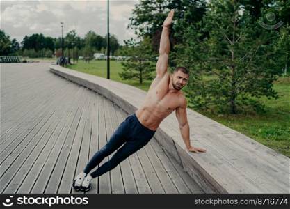 Athletic man stands in side plank pose, raises one arm, poses with shirtless torso, wears trousers and sneaks. Sportsman exercises in park on fresh air, has perfect fit muscles body. Healthy lifestyle