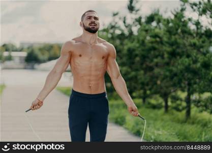 Athletic motivated man with naked torso concentrated on intense fitness training program jumps over skipping rope, poses in park has sporty body, breathes air deeply. Morning workout concept