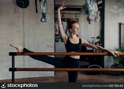 Athletic red haired woman in sleeveless black top tank and leggings stretching leg on ballet barre against sport equipment background of fitness studio, ballerina exercising in fitness studio. Young athletic red haired ballerina stretching leg on ballet barre in fitness studio