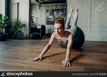 Athletic slender lady with red hair performing stretching exercises on top of big silver fitball in ambience of fitness studio background, being concentrated on exercise for strength and balance. Athletic slender lady with red hair performing streching exercises on big silver fitball during pilates workout