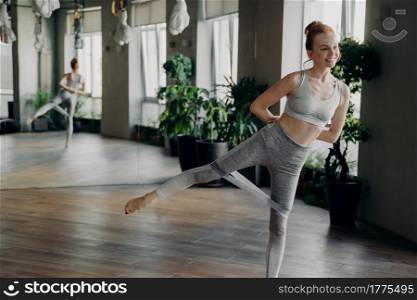 Athletic slender red haired woman during fitness workout with rubber resistance band, raises leg up for hip muscles activation, keeps hands behind. Concept of sport and active lifestyle concept. Athletic slender red haired woman during her fitness workout with rubber resistance band