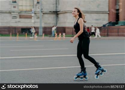 Athletic woman on roller skates moves actively poses on asphalt at street dressed in active wear has cheerful expression. Young fitness model skating in urban place. Sporty lifestyle rollerblading