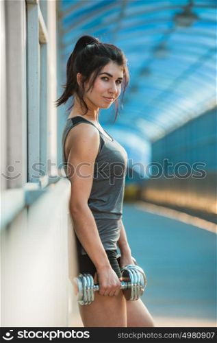 Athletic woman pumping up muscles with dumbbells outdoors in the city