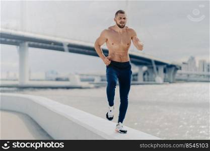 Athletic young bearded man goes in for sport has morning run breathes deeply prepares for running marathon. Motivated sportsman jogging by river has strong healthy body. Fitness and sport concept