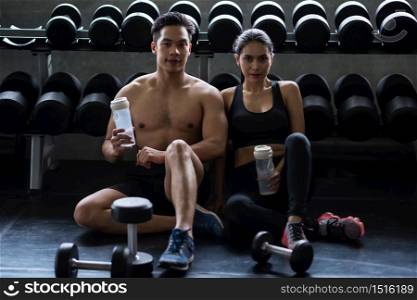 Athletic young muscular Asian couple with sexy muscle body break and drink water near dumbbell shelf after workout or exercise in fitness gym. Bodybuilding and healthy lifestyle concept.