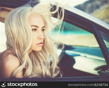 Atmospheric outdoors fashion portrait of beautiful blonde in car on the beach