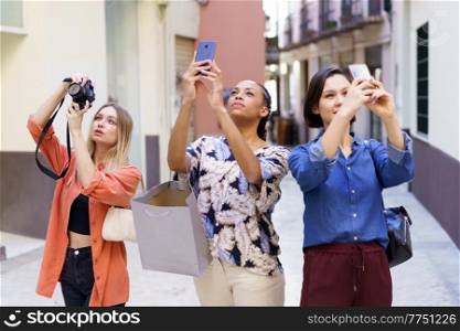 Attentive young multiracial female tourists in casual clothes taking photos on digital camera and smartphones while sightseeing city during holiday together. Stylish young diverse women taking photos during vacation in old city
