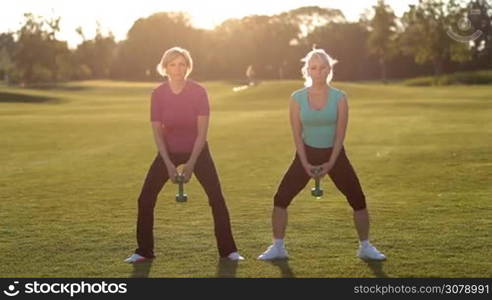 Attracive fitness senior females in sporstwear working out with dumbbells, doing crossfit squatting on park lawn in glow of beautiful sunset. Sporty adult women in great shape exercising outdoors, performing squats with light dumbbells.