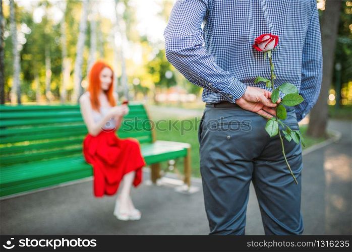 Attracrive woman sits on a bench in park, man with rose stands against. Romantic date of love couple