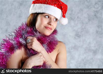 Attractive 20-25 years brunette smiling topless headshot woman in Santa hat studio on grey.