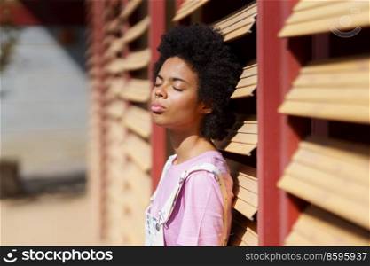 Attractive African American female with Afro hairstyle standing with closed eyes under bright sunlight near wooden wall on sunny street. Dreamy black woman enjoying sunny day