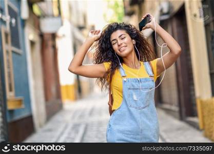 Attractive African girl listening to music with earphones outdoors. Arab woman in casual clothes with curly hairstyle in urban background with closed eyes.. Attractive African girl listening to music with earphones outdoors.