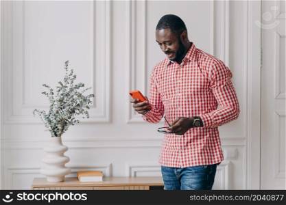 Attractive Afro American businessman in casual wear holding smartphone trying to answer important call. Confident mixed race man entrepreneur works remotely from modern home office. Freelance concept. Attractive Aframerican businessman in casual wear holding smartphone trying to answer important call