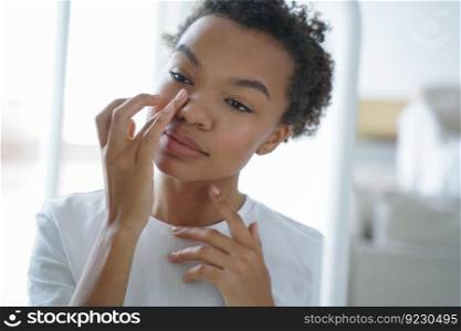 Attractive afro girl is applying cream touching her face. Femininity and grooming concept. Mirror reflection of young latin woman. Skin cleansing. Daily routine, cosmetics and hygiene of teenage girl.. Attractive afro girl is applying cream touching her face. Mirror reflection of young latin woman.