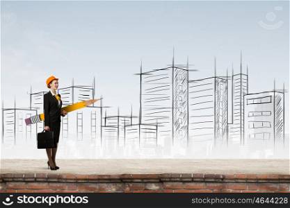 Attractive architect designer. Young female engineer in hard hat with big pencil and sketches at background