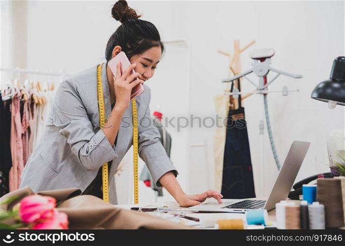 Attractive Asian female fashion designer working in clothing shop studio and using mobile phone to contact customer. Woman receiving order from incoming call. Tailor sewing. People lifestyle concept