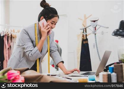 Attractive Asian female fashion designer working in clothing shop studio and using mobile phone to contact customer. Woman receiving order from incoming call. Tailor sewing. People lifestyle concept