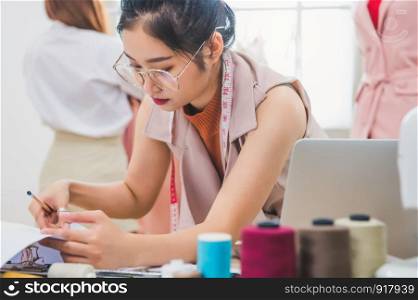 Attractive Asian female fashion designer working in clothing shop with customer background. Stylish fashionista woman creating new cloth design collection. Tailor and sewing. People lifestyle concept