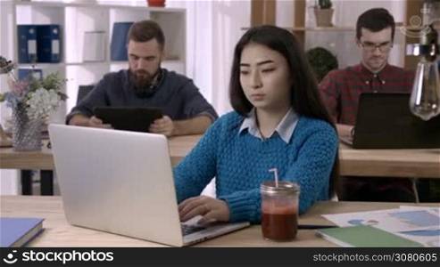 Attractive asian girl working with documents and laptop in open space office with coworkers using digital devices on background. Young female freelancer drinking healthy berry smoothie at workplace.