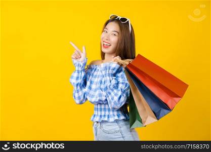 Attractive Asian smiling young woman Carrying a shopping coloful bag and pointing the sale advertisement on isolated yellow color background, copy space and studio, black friday season sale concept.