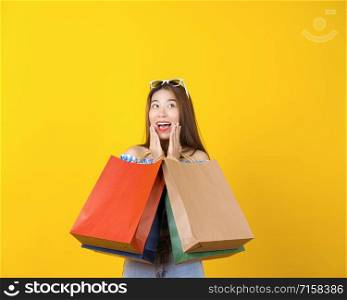 Attractive Asian smiling young woman Carrying a shopping coloful bag and pointing the sale advertisement on isolated yellow color background, copy space and studio, black friday season sale concept.