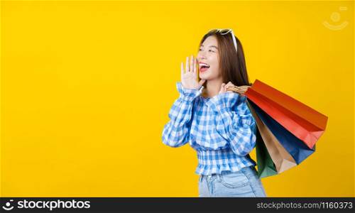 Attractive Asian smiling young woman Carrying a shopping coloful bag on aisolated yellow color background, copy space and studio, black friday season sale concept.