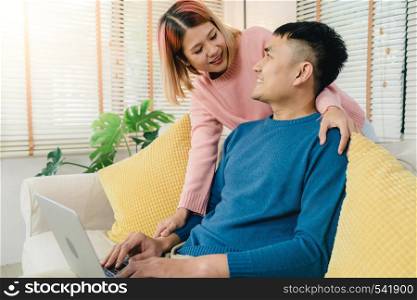 Attractive Asian sweet couple using computer or laptop while lying on the sofa when relax in their living room at home. Husband and his wife using relax and romantic time at home concept.
