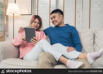 Attractive Asian sweet couple using tablet while lying on the sofa when relax in their living room at home. Husband and his wife using relax and romantic time at home concept.