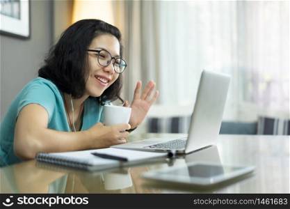 Attractive Asian woman making video call with laptop computer and waving hand. Young female in blue shirt talking with family and people via internet technology at home. Online E-learning and studying