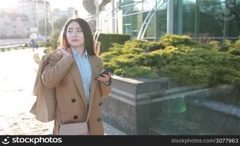 Attractive asian woman with shopping bags browsing the internet with smartphone while walking on cobblestone street at sunset. Cheerful relaxed girl socializing on mobile phone via internet in social media after good day shopping. Slow motion.