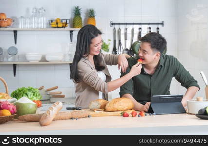 Attractive Asian young sweet couple cooking together in home kitchen. Beautiful wife smiling preparing breakfast and feeding strawberry to her husband while he working on tablet. Family joyful moment