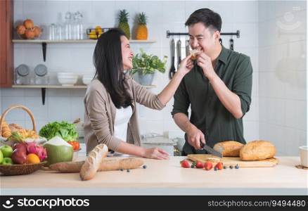 Attractive Asian young sweet couple cooking together in home kitchen. Beautiful woman smiling feeding her handsome man with bread while preparing meal, fresh fruits for breakfast. Family joyful moment