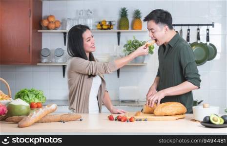 Attractive Asian young sweet couple cooking together in home kitchen. Beautiful woman smiling feeding her handsome man with bread while preparing meal, fresh fruits for breakfast. Family joyful moment