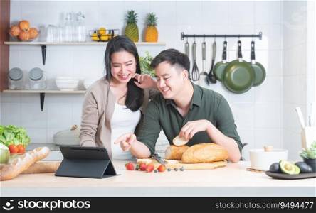 Attractive Asian young sweet couple looking recipe by using tablet at home kitchen, smiling, enjoy cooking preparing healthy breakfast meal with bread and fresh fruits together. Family, food concept