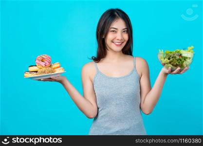 Attractive Asian young woman holding and choosing between disk of donuts or vegetable salad in glasses bowl on isolated blue color background, weight loss and avoid junk food for dieting and Healthy